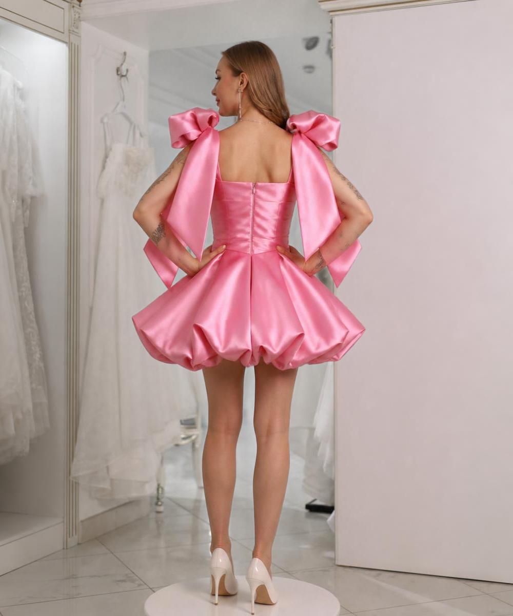 Pink satin mini dress with a voluminous bubble skirt and large 3D bows on the shoulder straps, worn with white heels.