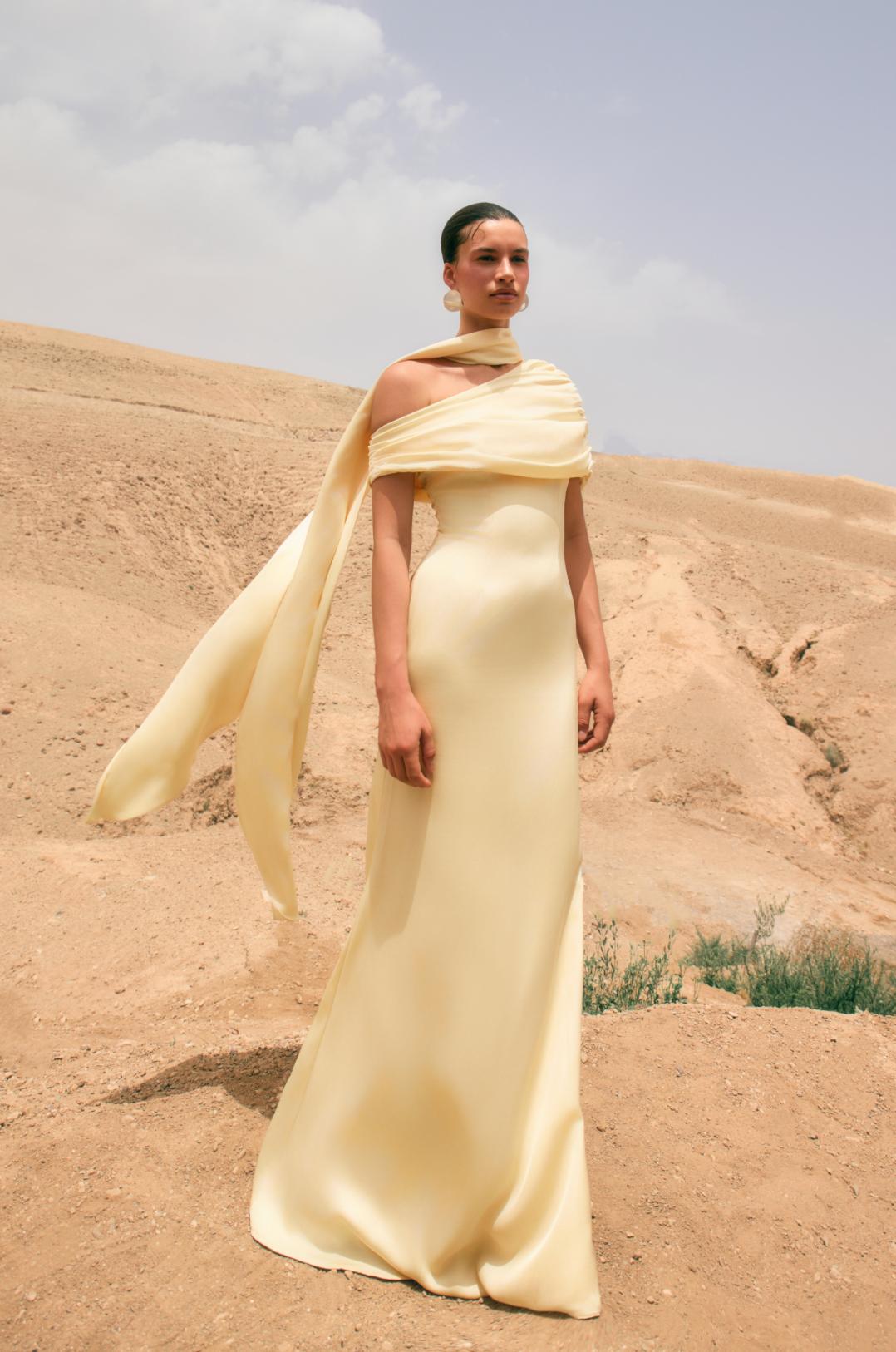 Model wearing a pale yellow satin Kulture dress with a gathered Bardot neckline and flowing A-line silhouette in a desert.