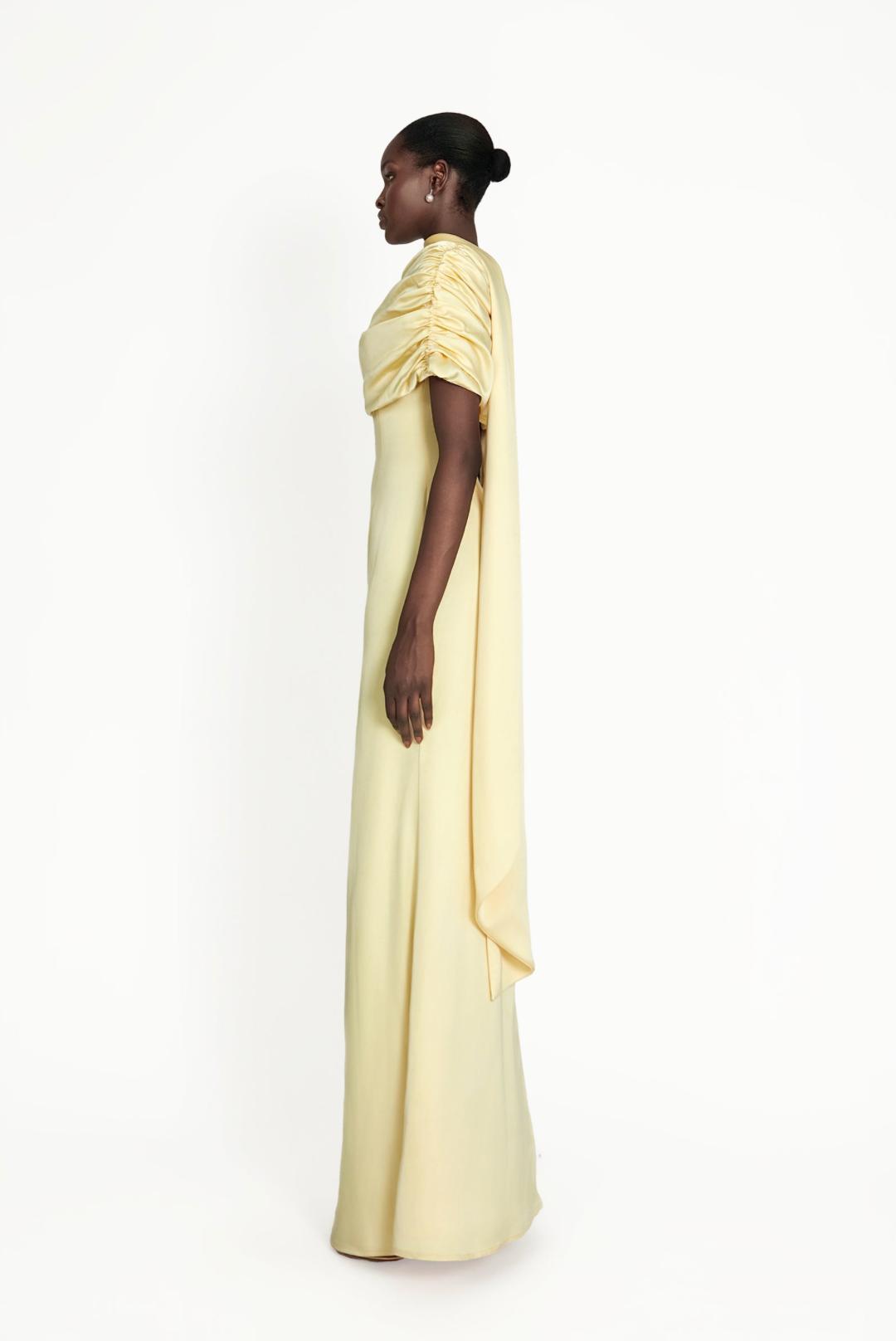 Model wearing a pale yellow satin Kulture dress with a gathered Bardot neckline and flowing A-line silhouette.