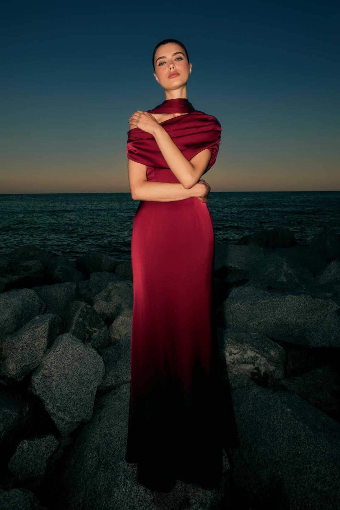 Woman wearing a deep red satin Kulture dress with a gathered Bardot neckline, standing on rocks by the ocean at dusk.