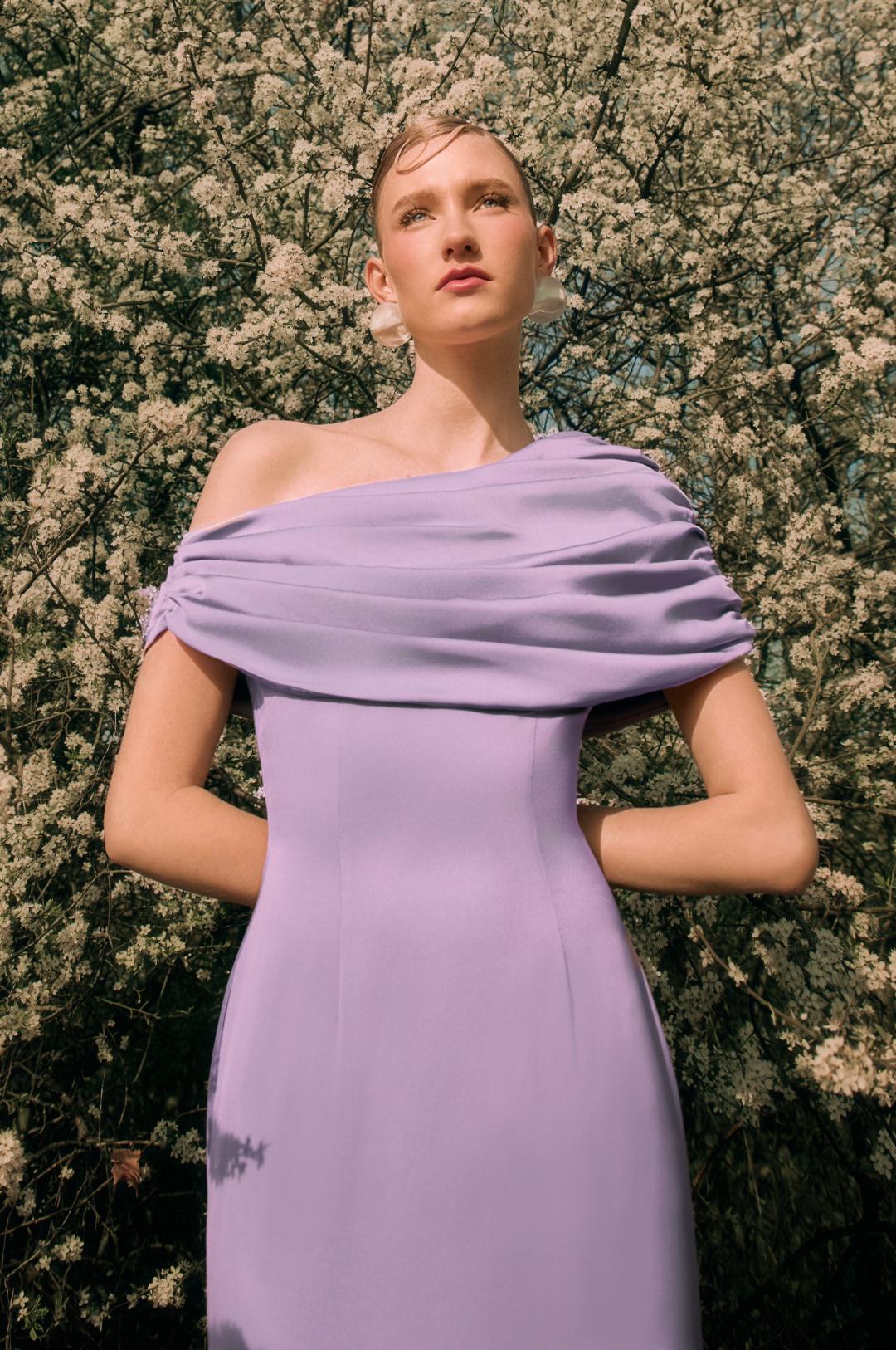 Lavender satin Kulture dress with a gathered Bardot neckline and A-line silhouette standing before white blossoms.
