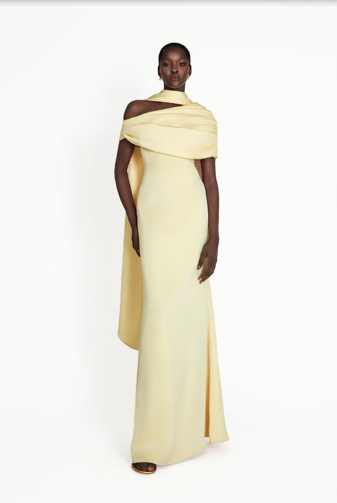 Model wearing a pale yellow satin Kulture dress with a gathered Bardot neckline and flowing A-line silhouette.