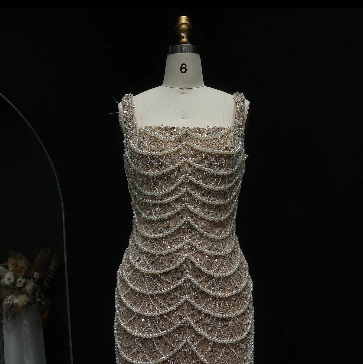 Nude sleeveless sheath gown featuring a scoop neckline, intricate pearl draping, and sparkling beadwork on a mannequin.