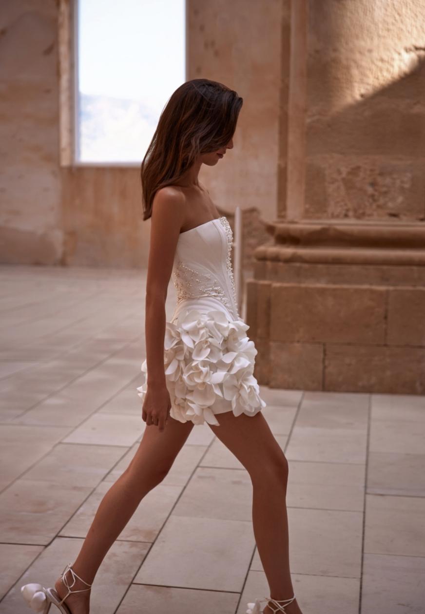 White strapless bridal mini dress with 3D floral appliqués, pearl beading, and a structured bodice in a stone-walled setting.
