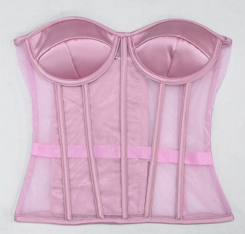 The Lilah light pink structured corset bustier with satin-like cups, sheer mesh panels, vertical boning, and a matching pink waistband, shown flat against a light gray background.