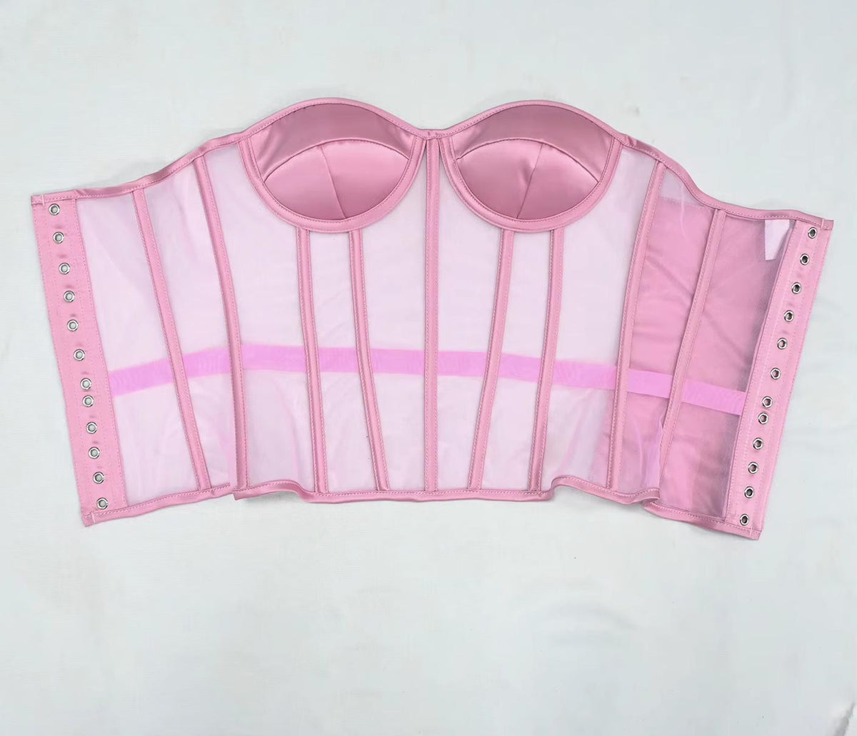The Lilah  pink structured bustier or corset-style top with sheer mesh panels, satin-like cup sections, vertical support panels, side straps, and metal eyelets, laid flat on a white background.