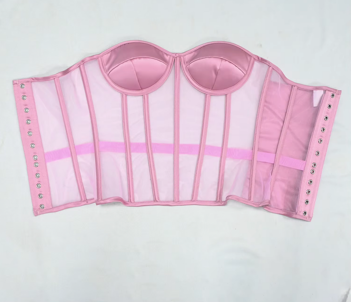 The Lilah  pink structured bustier or corset-style top with sheer mesh panels, satin-like cup sections, vertical support panels, side straps, and metal eyelets, laid flat on a white background.