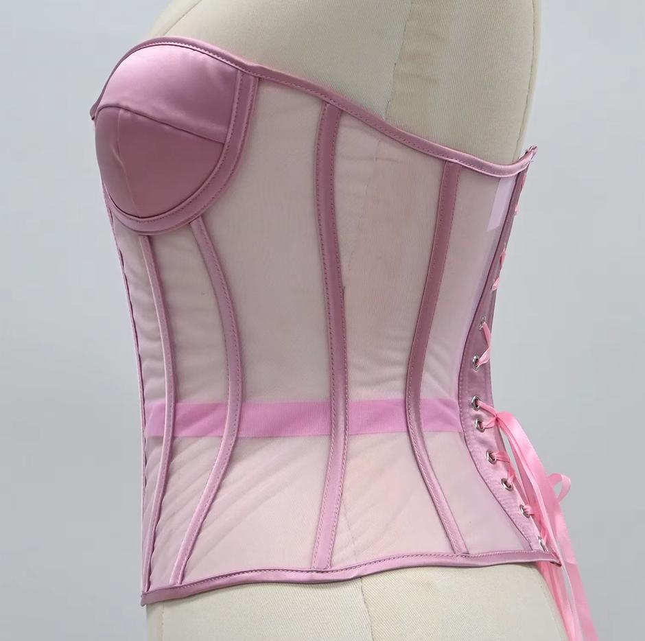 The Lilah light pink corset-style bustier top displayed on a cream mannequin torso against a plain light gray background. The garment features structured panels, sheer mesh sections, a padded bust area, side lacing, and a matching pink sash tied at the waist.