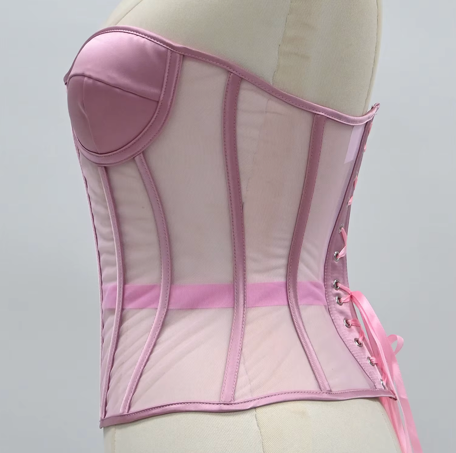 The Lilah light pink corset-style bustier top displayed on a cream mannequin torso against a plain light gray background. The garment features structured panels, sheer mesh sections, a padded bust area, side lacing, and a matching pink sash tied at the waist.