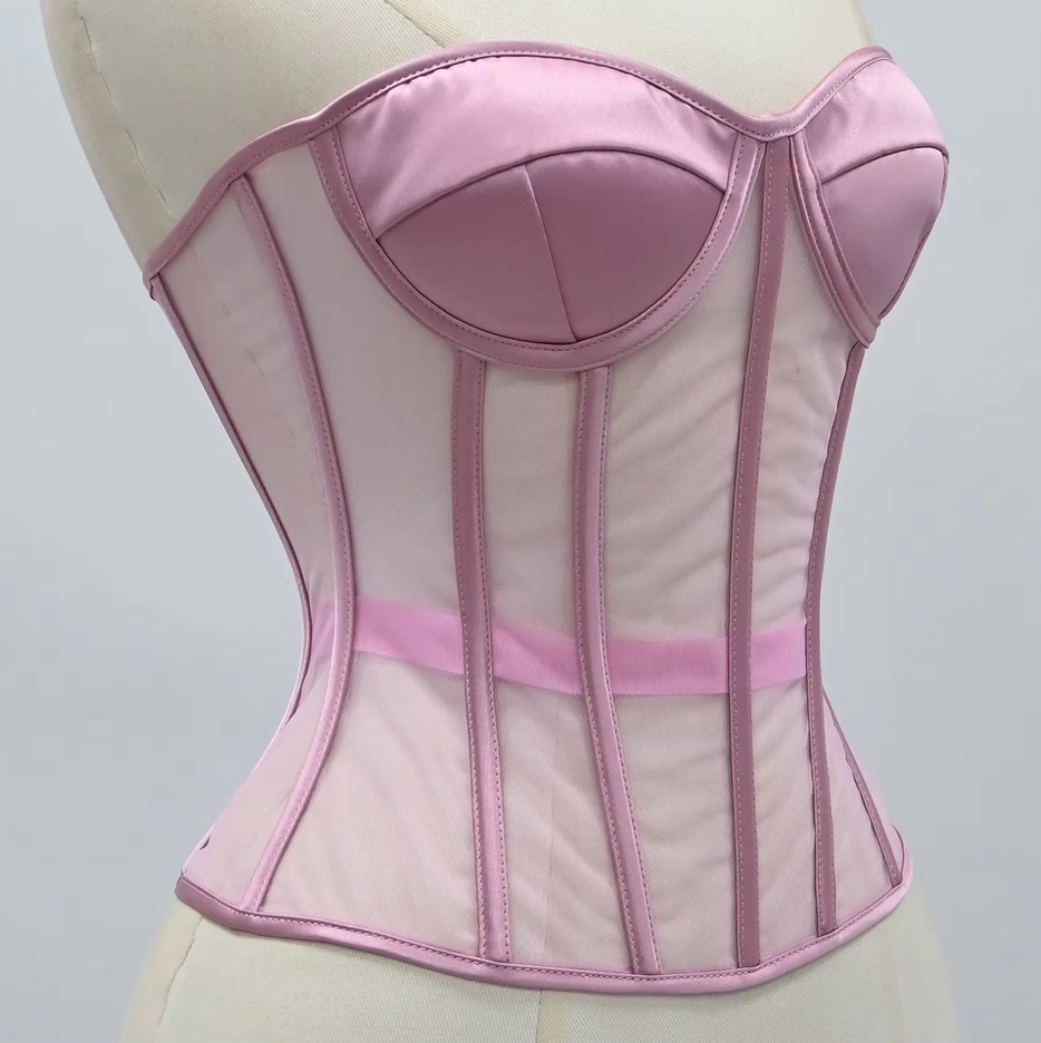 The Lilah light pink corset with satin-like panels and sheer mesh sections, displayed on a cream-colored dress form against a light gray background.