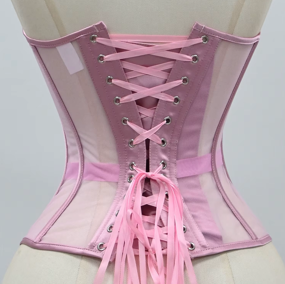 The Lilah pink corset-style harness with sheer mesh panels, structured fabric, and pink ribbon laces with metal eyelets, shown on a mannequin torso against a light gray background.