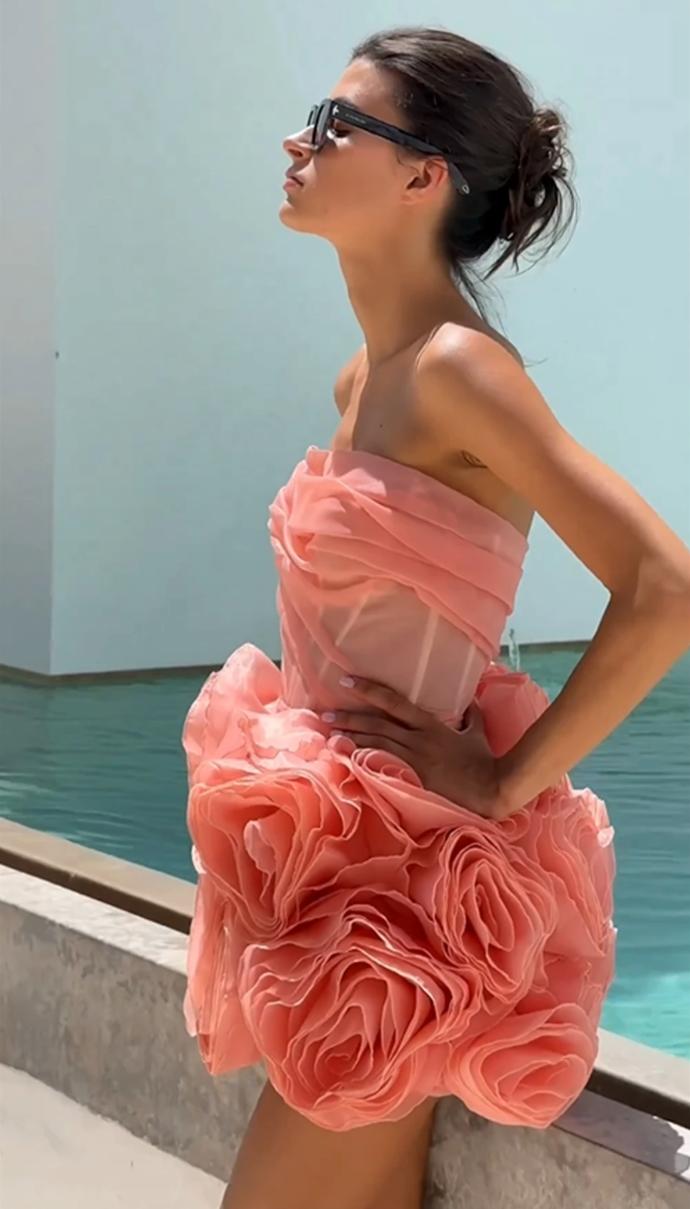 Model wearing a strapless peach mini dress with a corset bodice and 3D organza flower skirt by a pool.