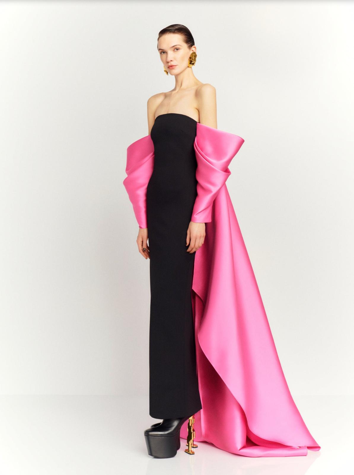 Model wearing a strapless black satin evening gown with dramatic pink draped sleeves and a long court train.