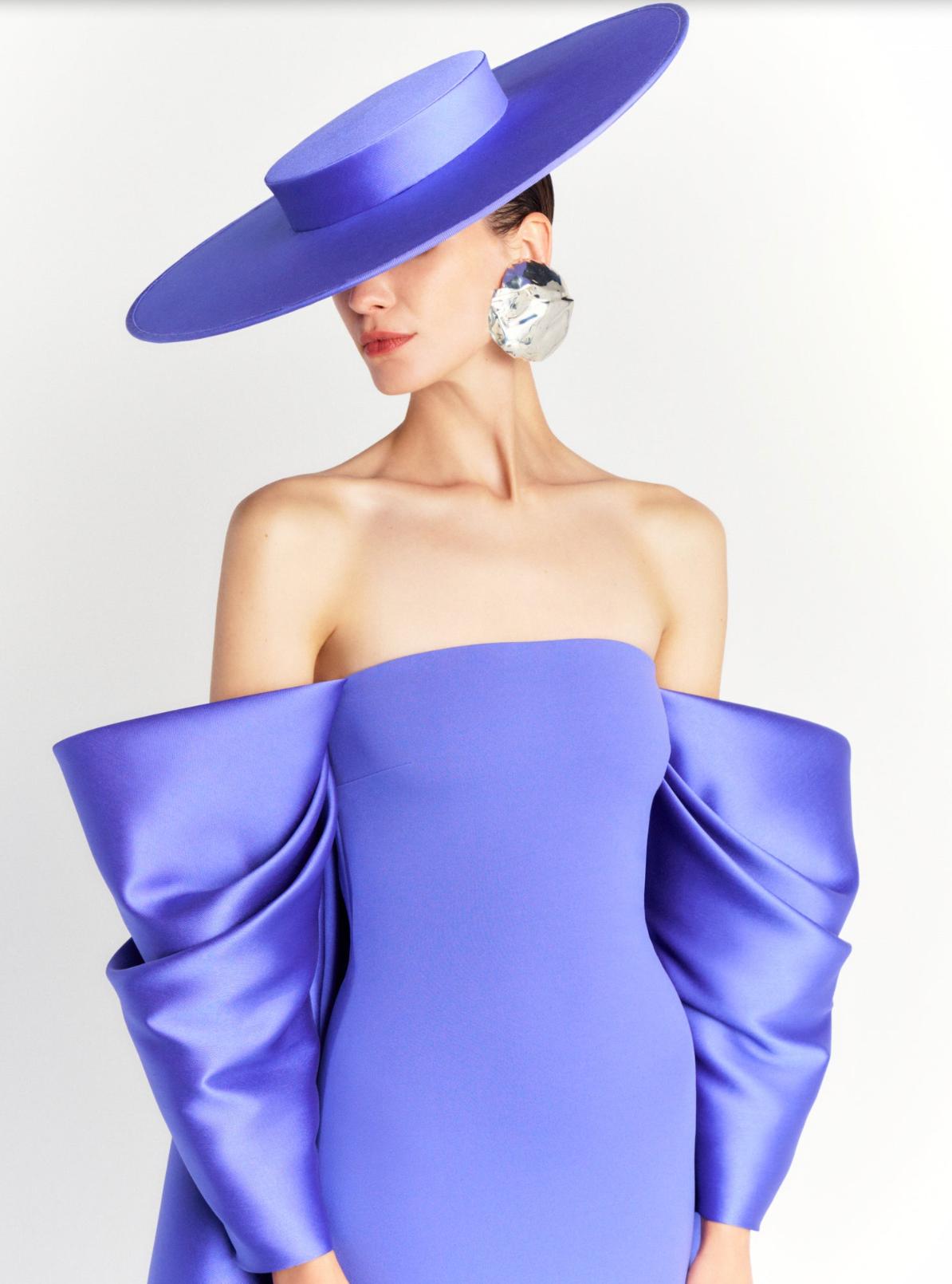 Model wearing a strapless royal blue satin evening gown with dramatic draped sleeves and a matching wide-brim hat.