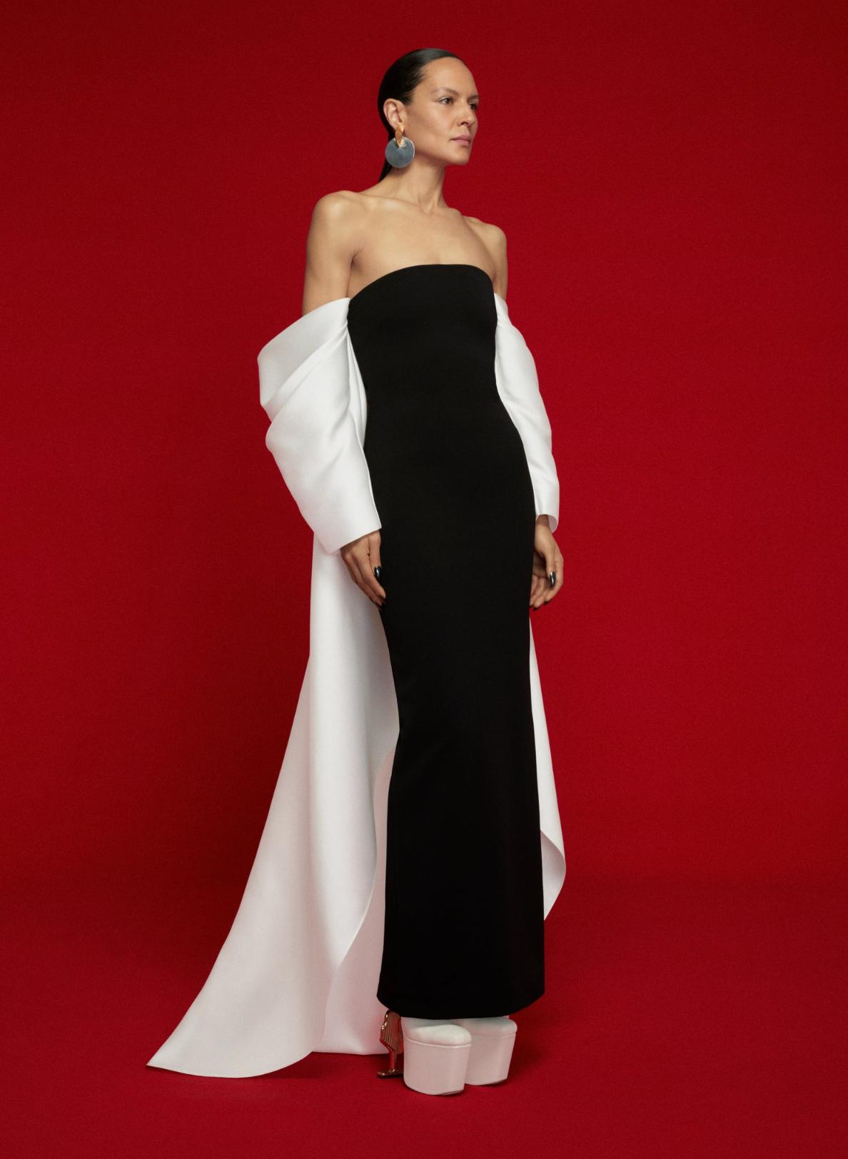 Strapless black satin evening gown with dramatic white off-shoulder sleeves and a long train against a red background.