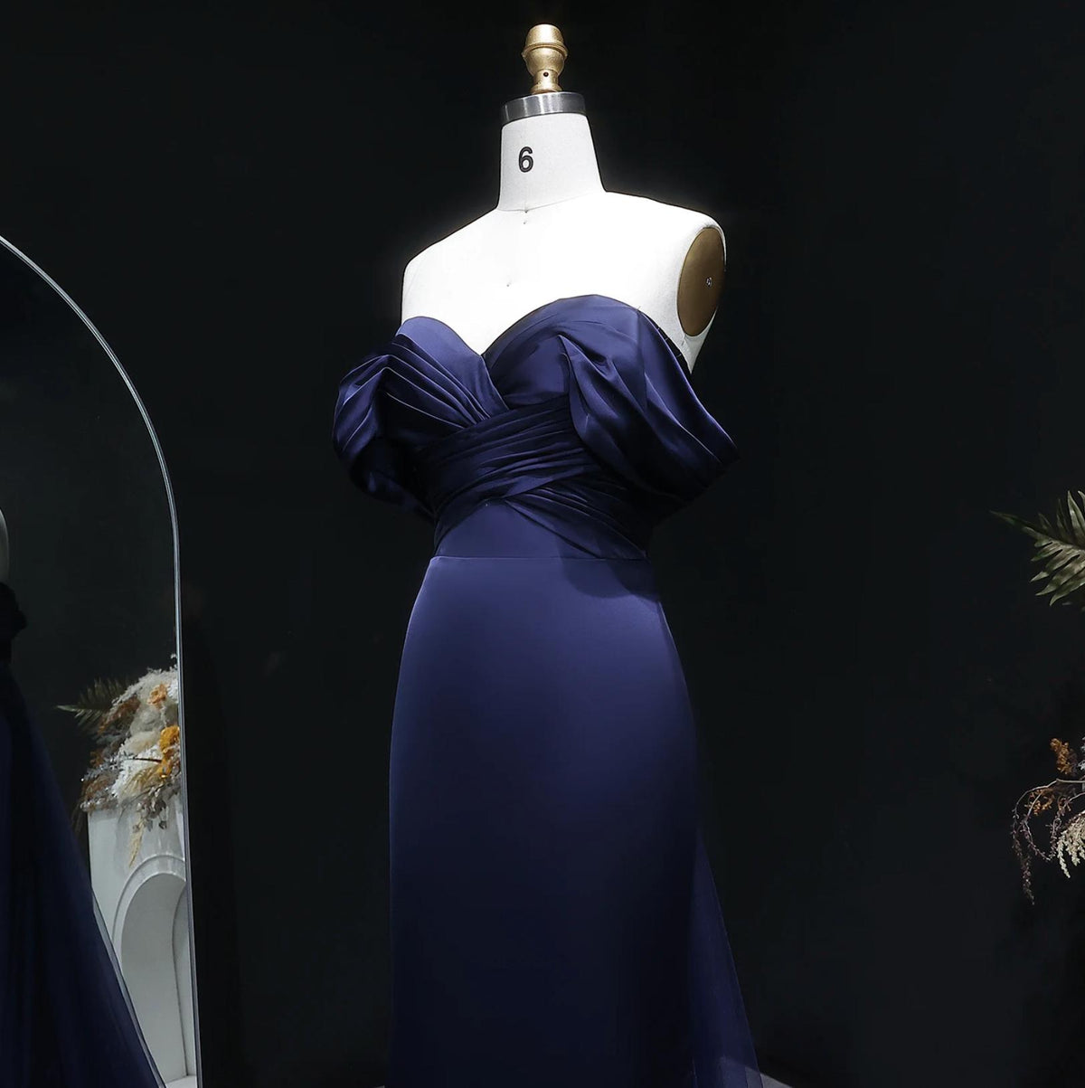 Navy blue satin mermaid evening gown with an off-shoulder pleated bodice on a dress form.