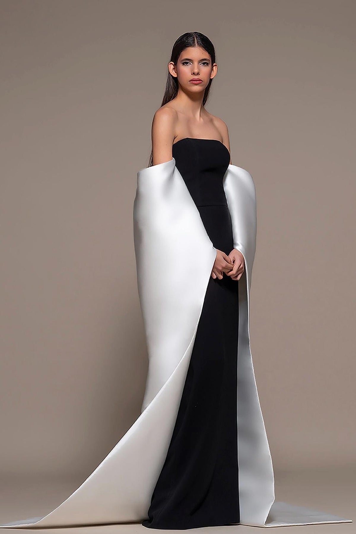 Model wearing a strapless black and white satin sheath evening gown with a sweeping train and structured draped sleeves.