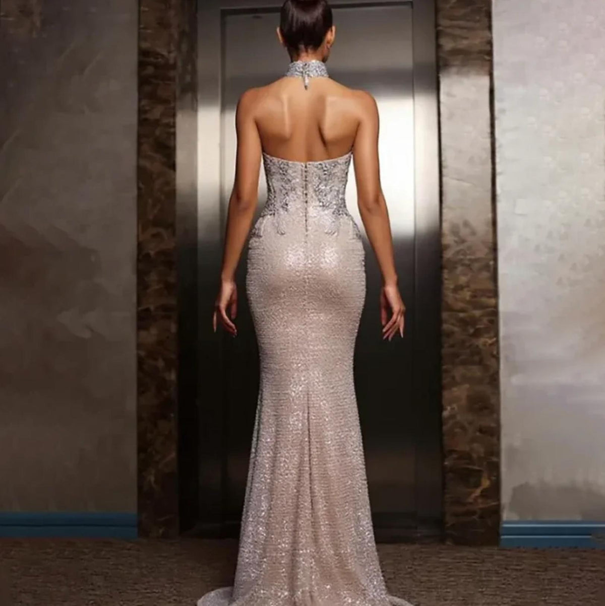 Backless silver floor-length gown with intricate beadwork, crystals, and a halter neckline in a fitted A-line silhouette.