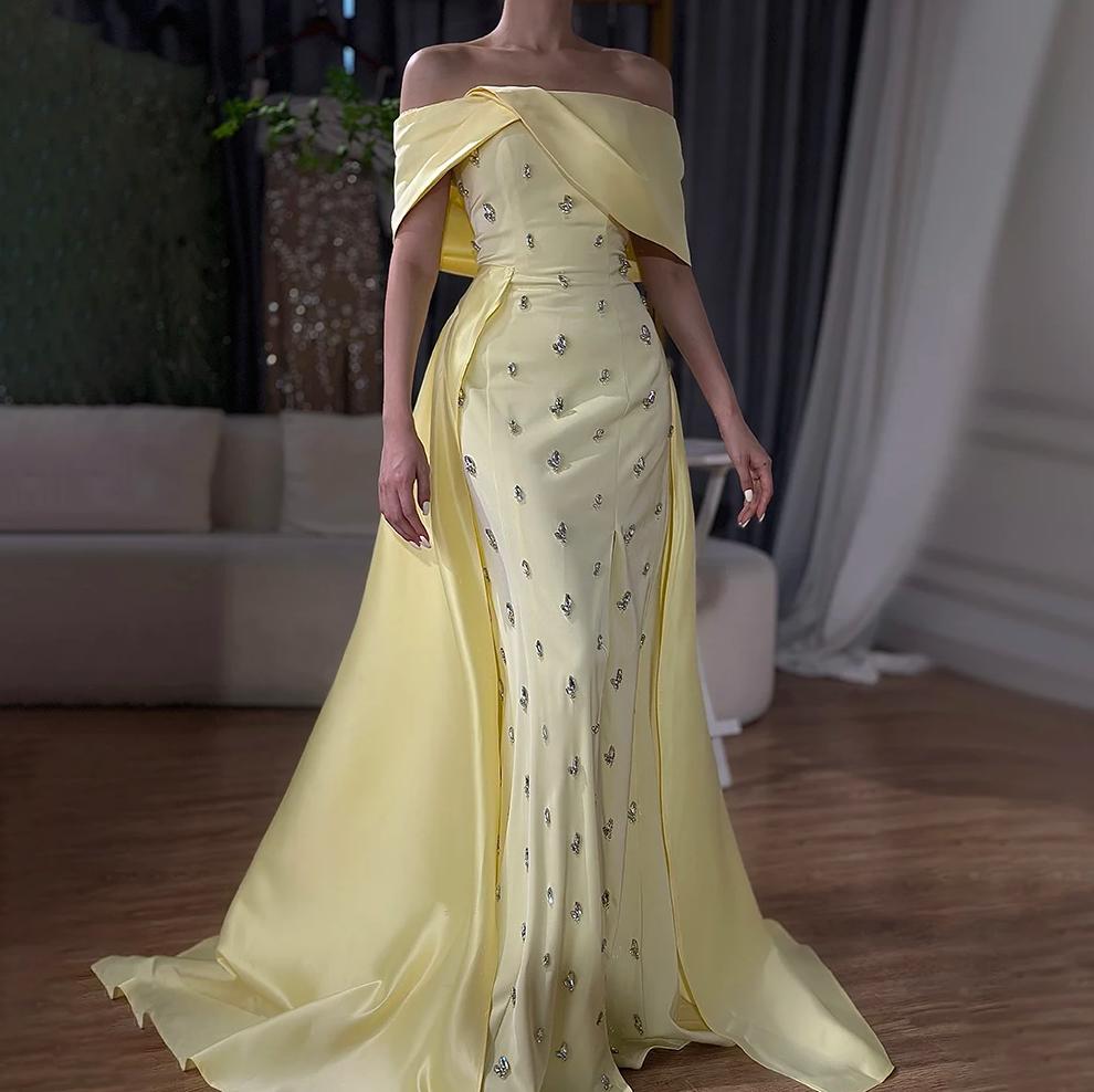 Pale yellow satin off-shoulder gown featuring a crystal-embellished bodice and a dramatic flowing overskirt.