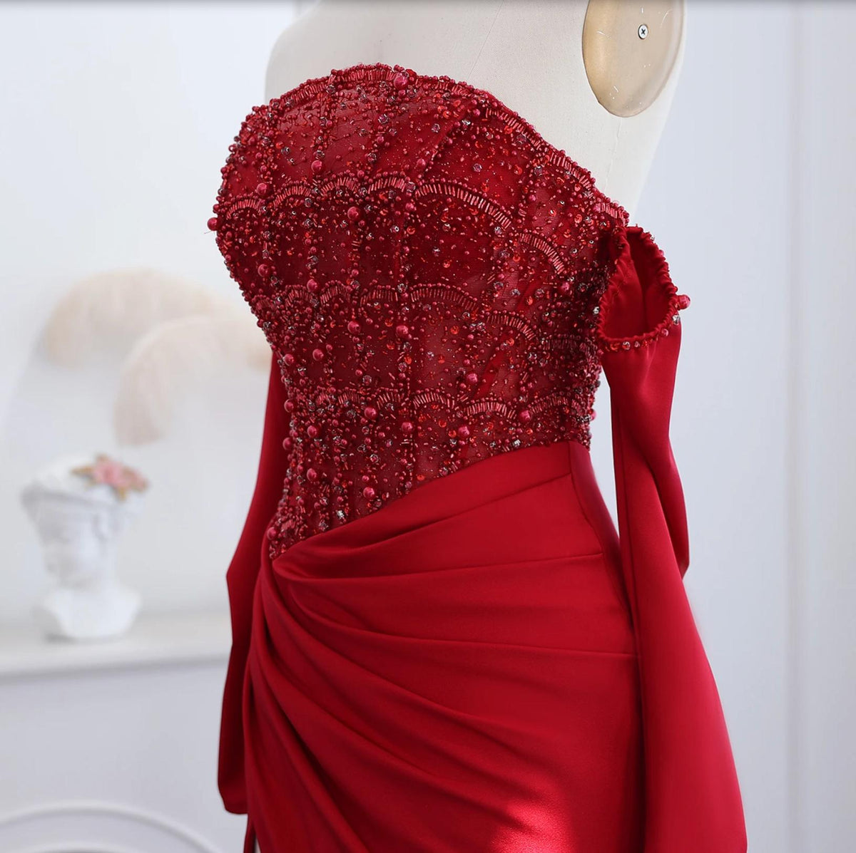 Red satin evening gown with an off-the-shoulder neckline, intricate crystal beading, and a draped bodice on a mannequin.