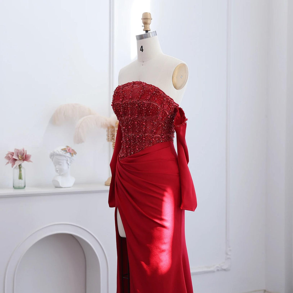 Red satin mermaid evening gown with a beaded bodice, off-the-shoulder neckline, and a high side slit on a mannequin.