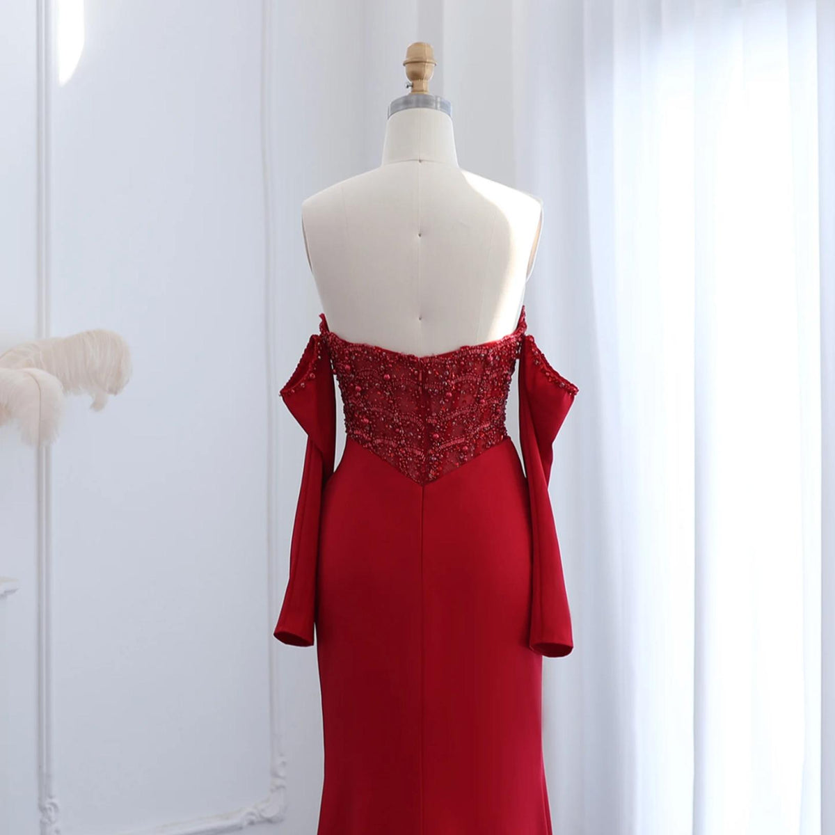 Red satin mermaid gown with off-the-shoulder sleeves and a beaded, crystal-embellished bodice on a mannequin.