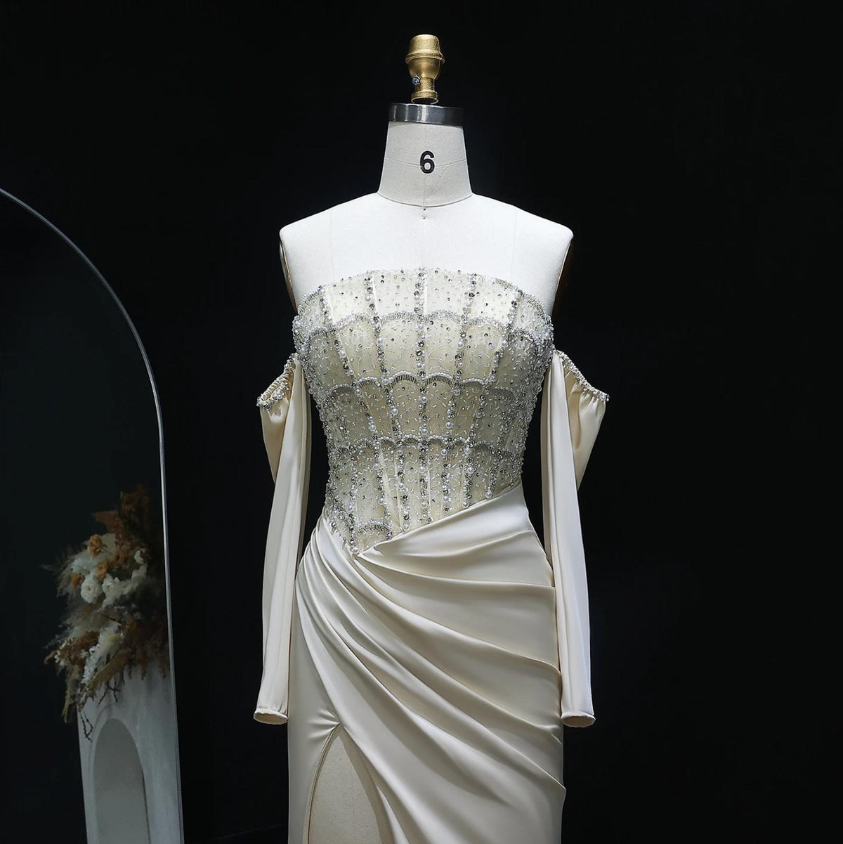 Cream satin evening gown with crystal-beaded bodice, off-the-shoulder sleeves, and a high slit on a dress form.