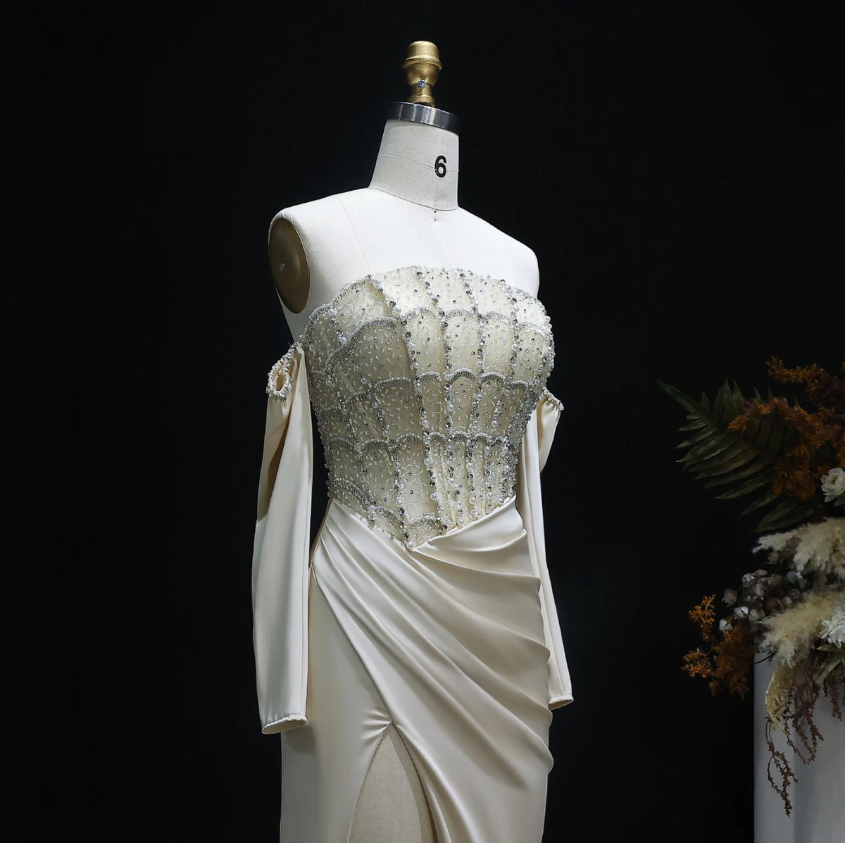 Cream satin evening gown with a beaded crystal corset bodice, off-shoulder sleeves, and a high-slit skirt on a mannequin.