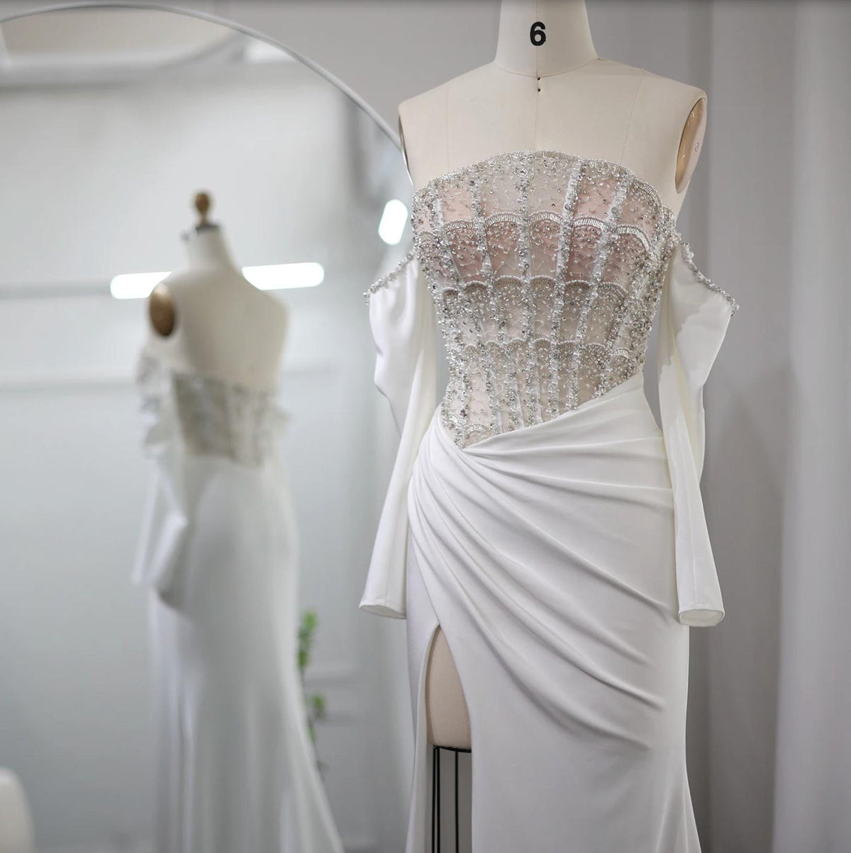 White satin mermaid gown with a beaded crystal bodice, off-the-shoulder sleeves, and a high slit on a mannequin.