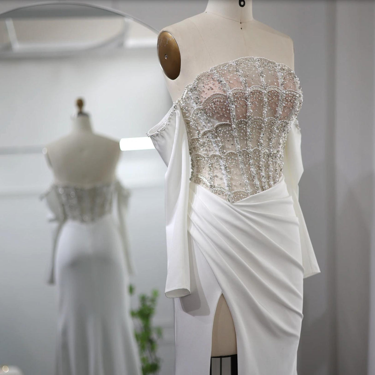 White satin evening gown with a beaded corset bodice, off-the-shoulder sleeves, and a high slit on a dress form.