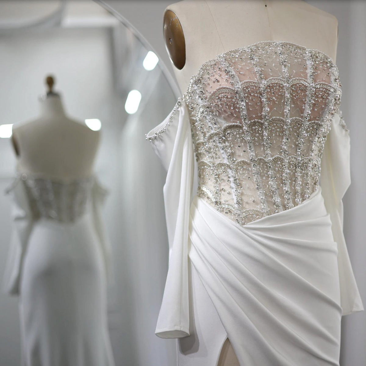 White satin evening gown featuring a crystal-beaded corset bodice, off-the-shoulder sleeves, and a high-slit skirt.