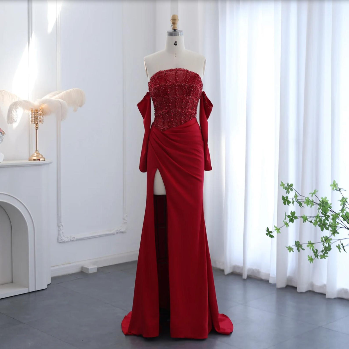 Red satin mermaid evening gown with a beaded corset bodice, off-the-shoulder sleeves, and a high front slit on a mannequin.