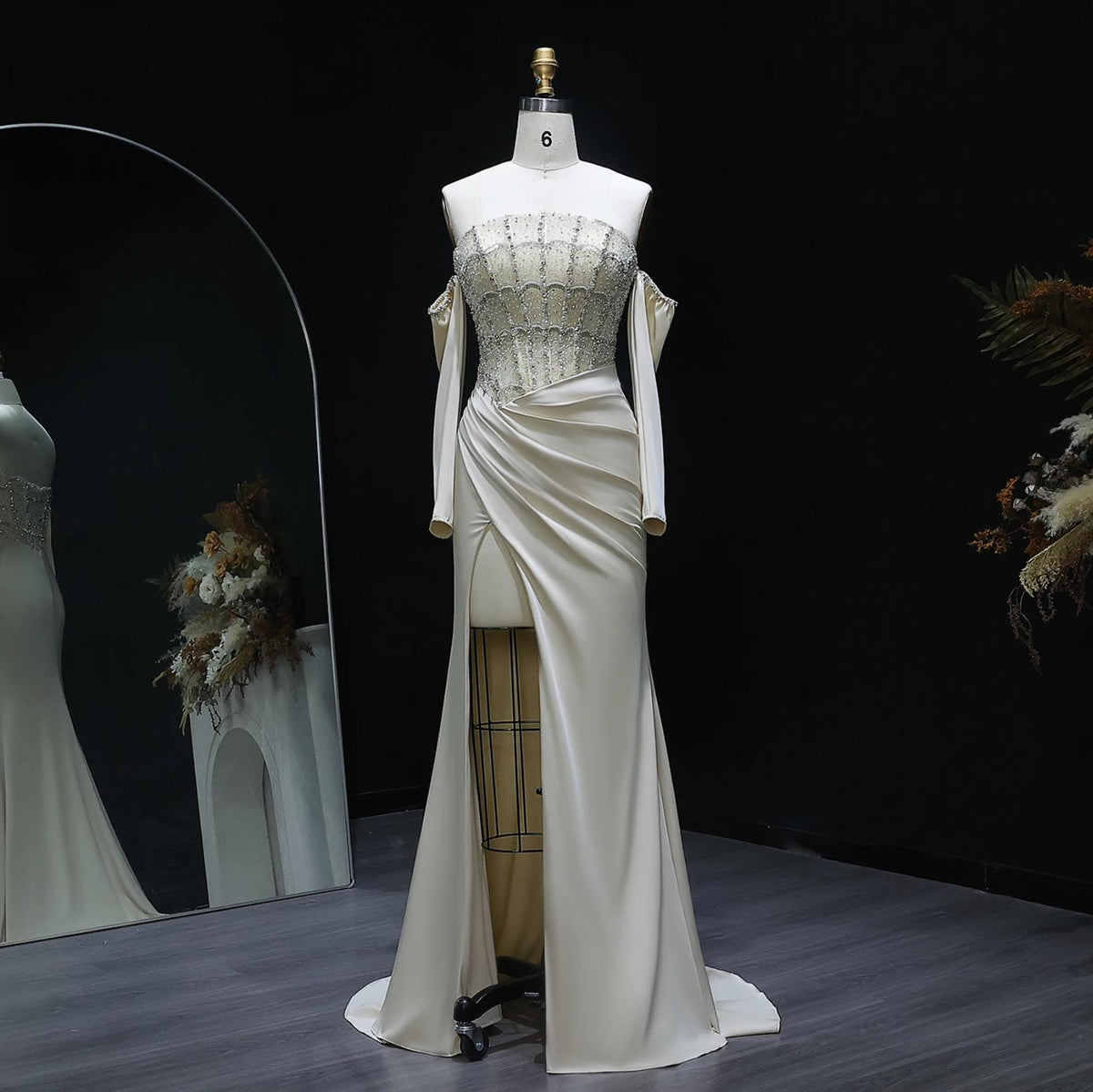 Off-the-shoulder cream satin evening gown with a crystal-beaded bodice, long sleeves, and a high-slit A-line skirt.