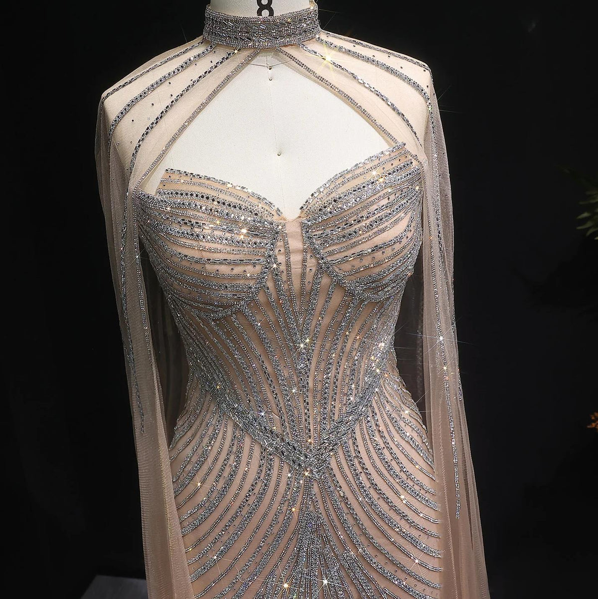Beaded mermaid gown with crystal-embellished tulle, high neckline, and dramatic cape sleeves on a mannequin.