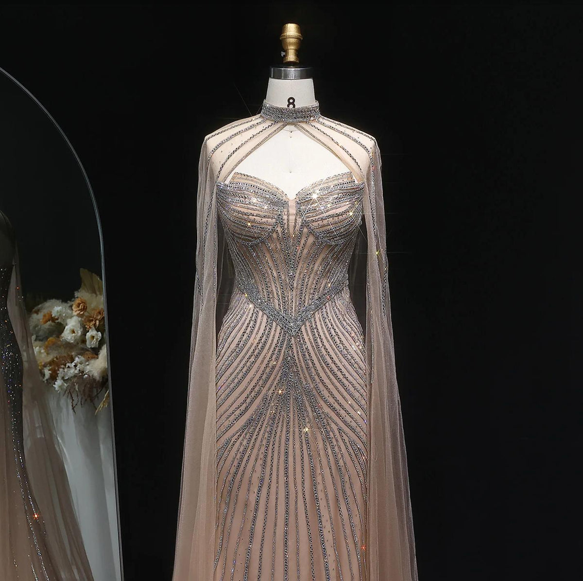 Beaded mermaid gown in nude tulle with crystal embellishments, high neckline, and dramatic sheer cape sleeves.