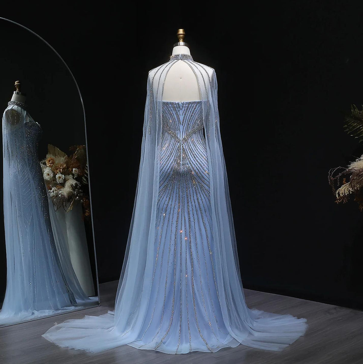 Blue tulle mermaid gown with crystal beading, dramatic cape sleeves, and a sweeping train on a mannequin.