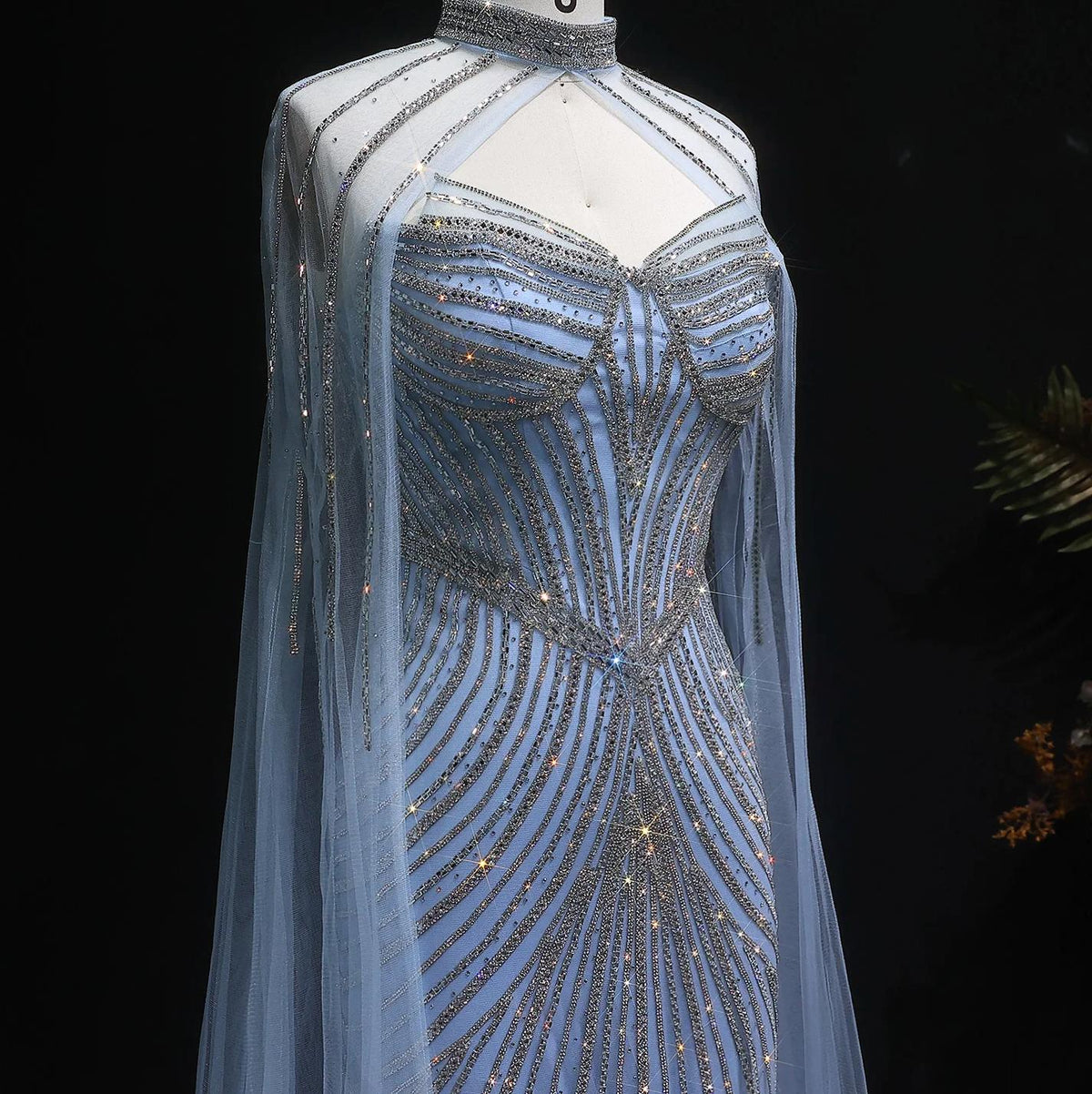 Ethereal blue tulle mermaid gown featuring intricate silver crystal beading, a high neckline, and dramatic cape sleeves.