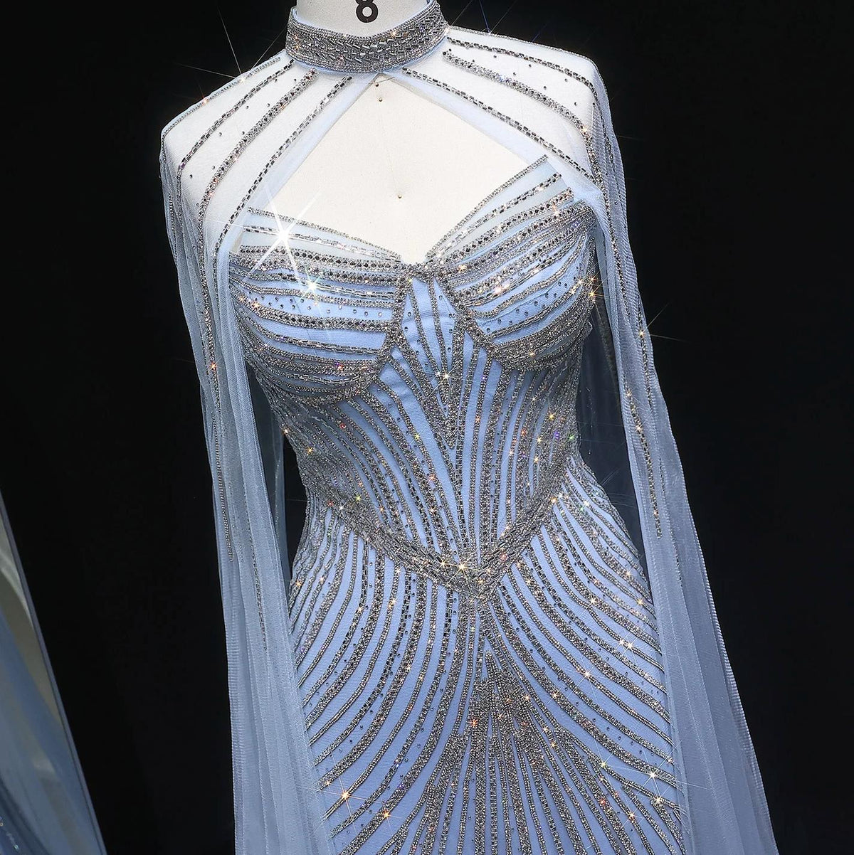 Blue tulle mermaid gown featuring intricate crystal beading, a high neck, and dramatic cape sleeves on a mannequin.