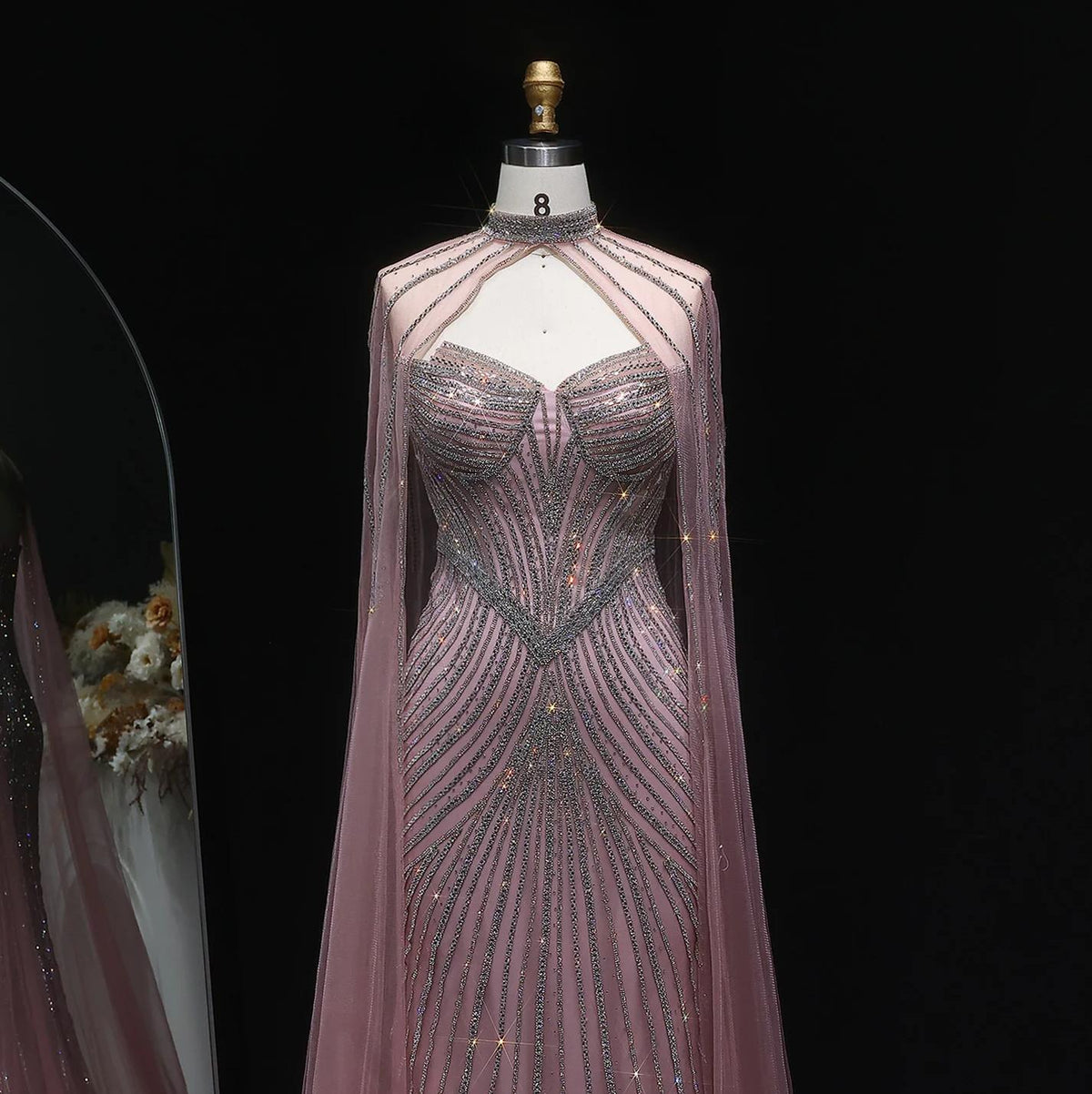Dusty pink mermaid gown with crystal beading, high neck, and dramatic tulle cape sleeves on a dress form.
