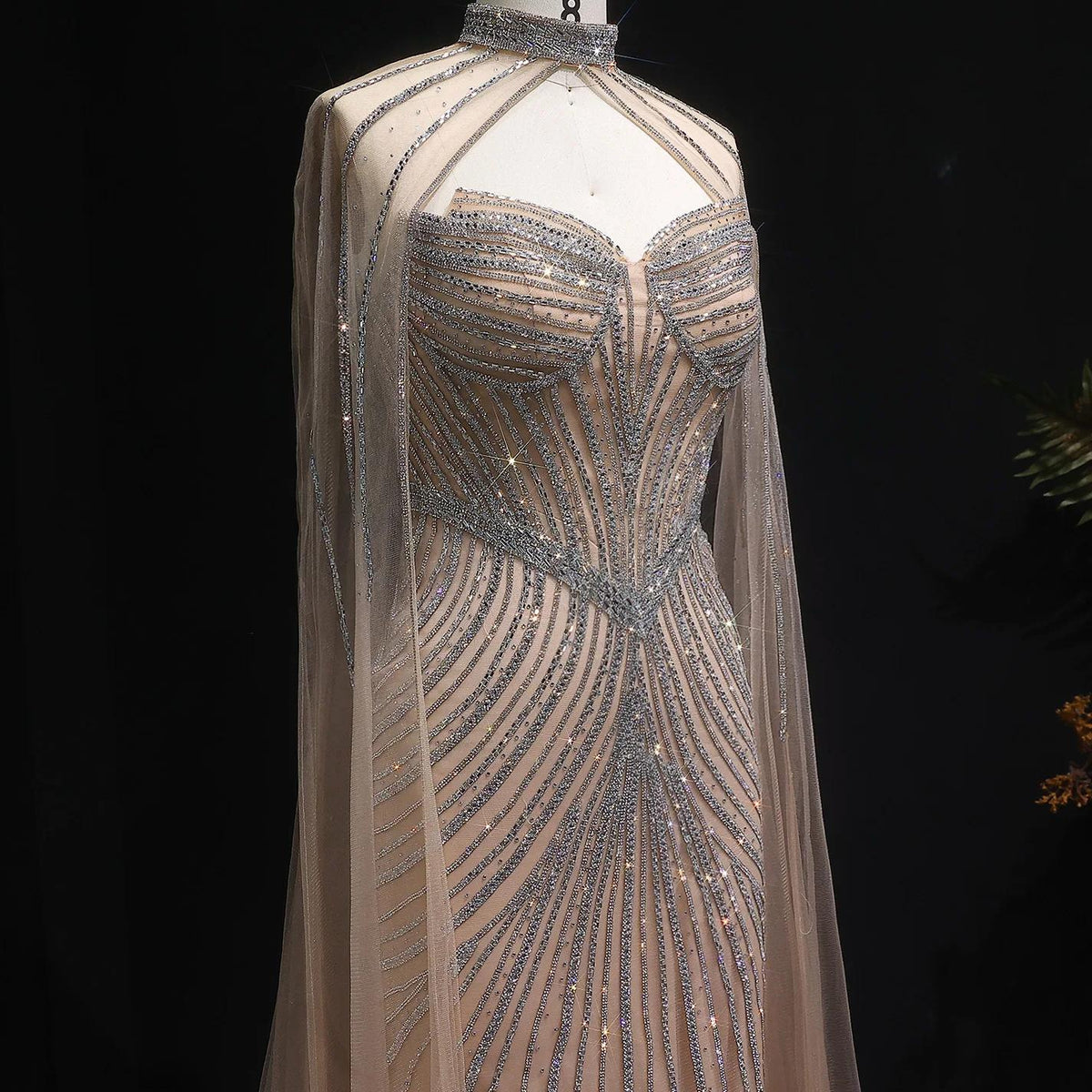 Beaded mermaid gown with crystal embellishments, sheer tulle cape sleeves, and a high-neck bodice on a mannequin.