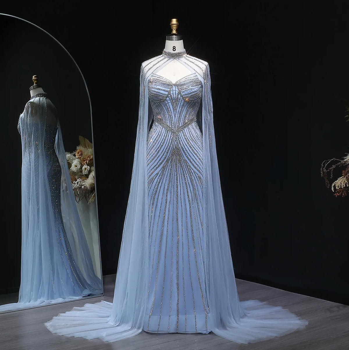Blue tulle mermaid gown with crystal beading, high neck, and dramatic cape sleeves on a mannequin.