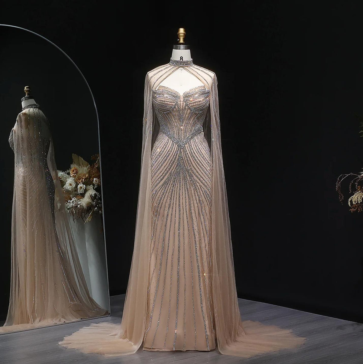 Beige mermaid gown with crystal beading, sheer cape sleeves, and a dramatic tulle sweep train on a mannequin.