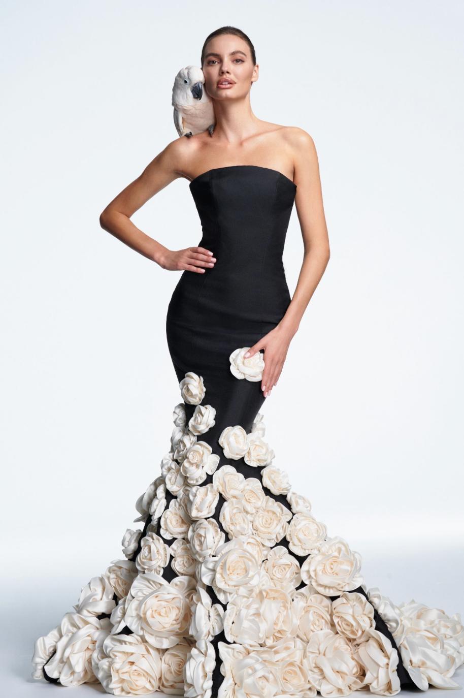 The Persia strapless black mermaid gown with a dramatic train covered in large cream-colored fabric flowers, posed against a clean white studio background.