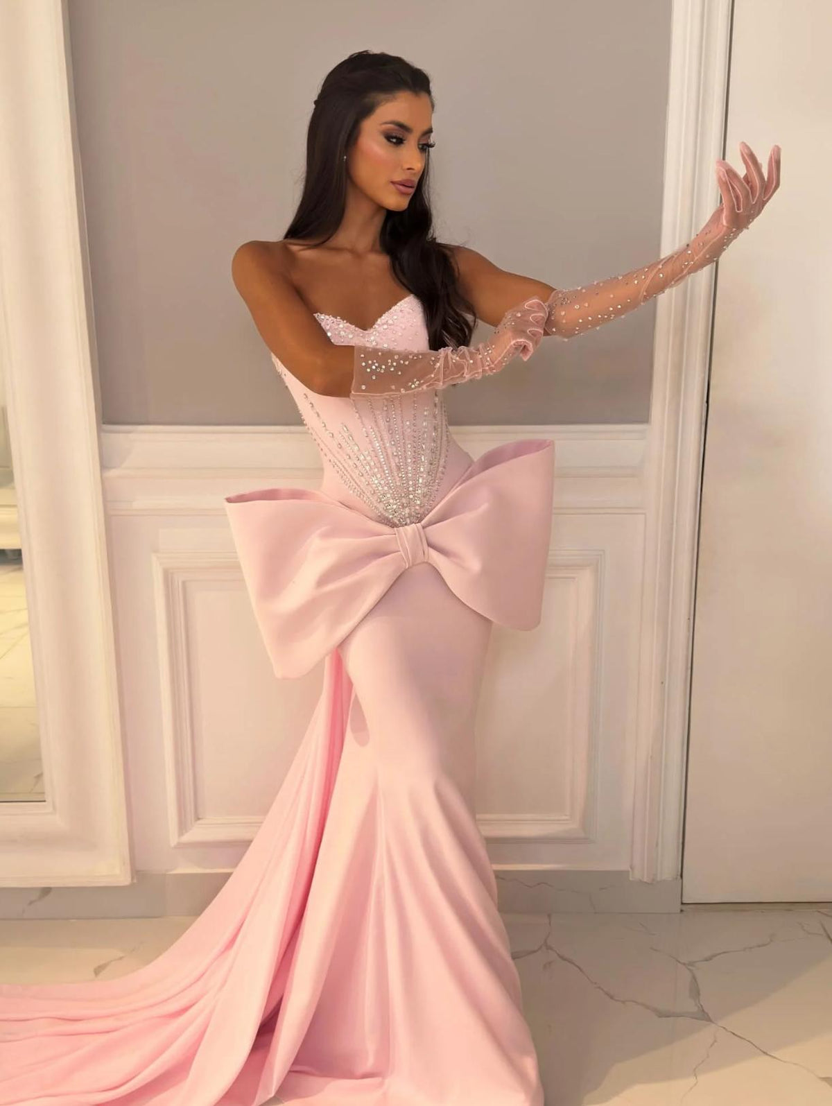 Pink strapless mermaid evening gown with a beaded corset bodice, large 3D waist bow, and matching embellished sheer gloves.