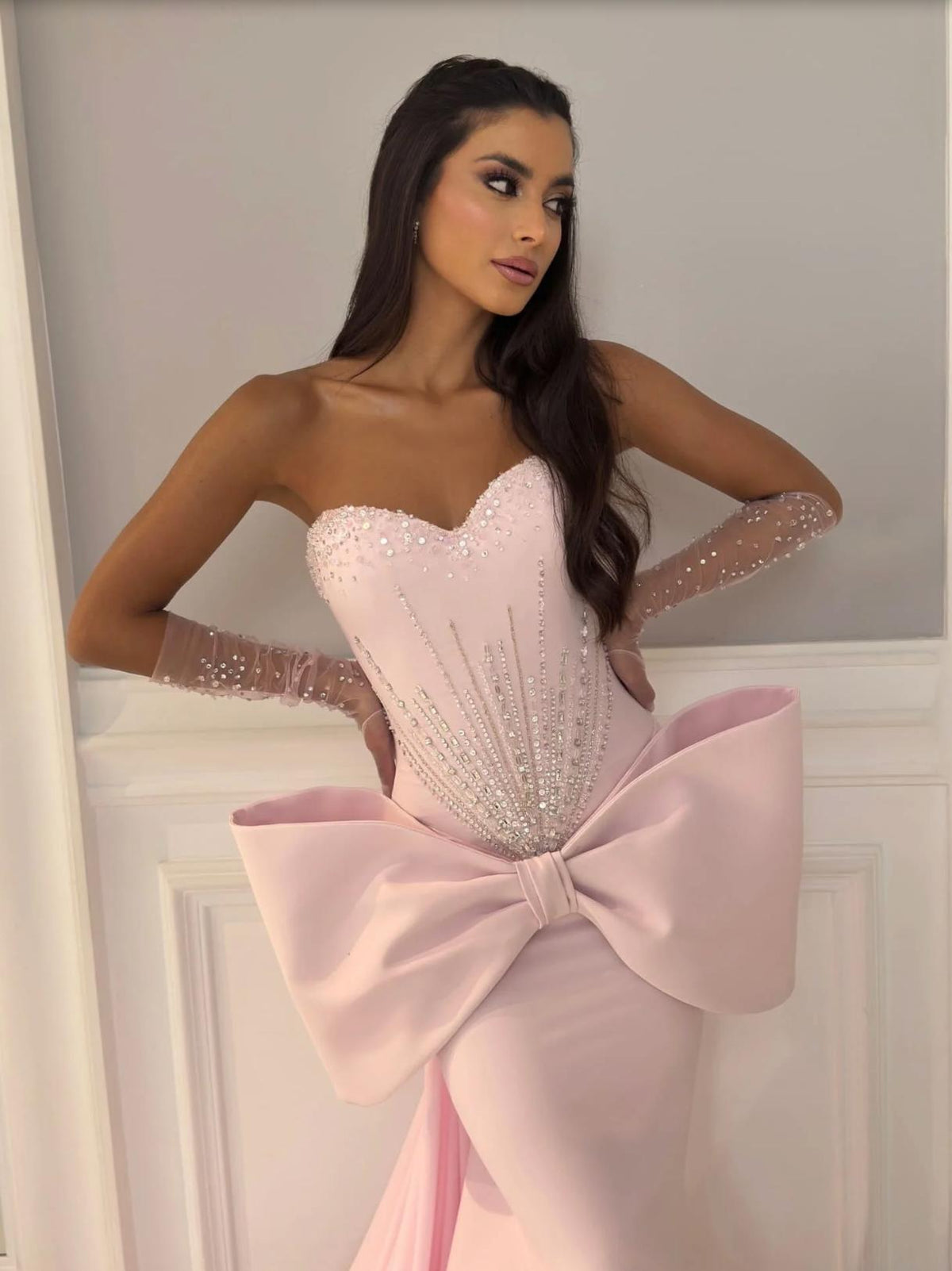 Strapless pink mermaid evening gown with a beaded corset bodice, dramatic 3D bow, and sheer embellished opera gloves.
