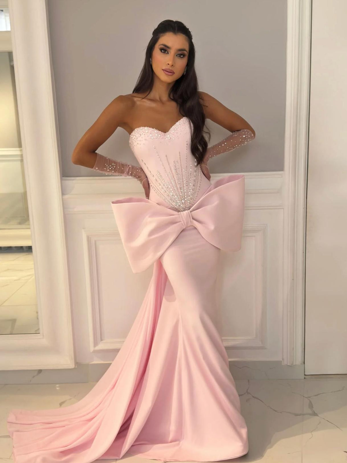 Pink strapless mermaid evening gown with a beaded corset bodice, dramatic 3D bow, and matching sheer embellished gloves.