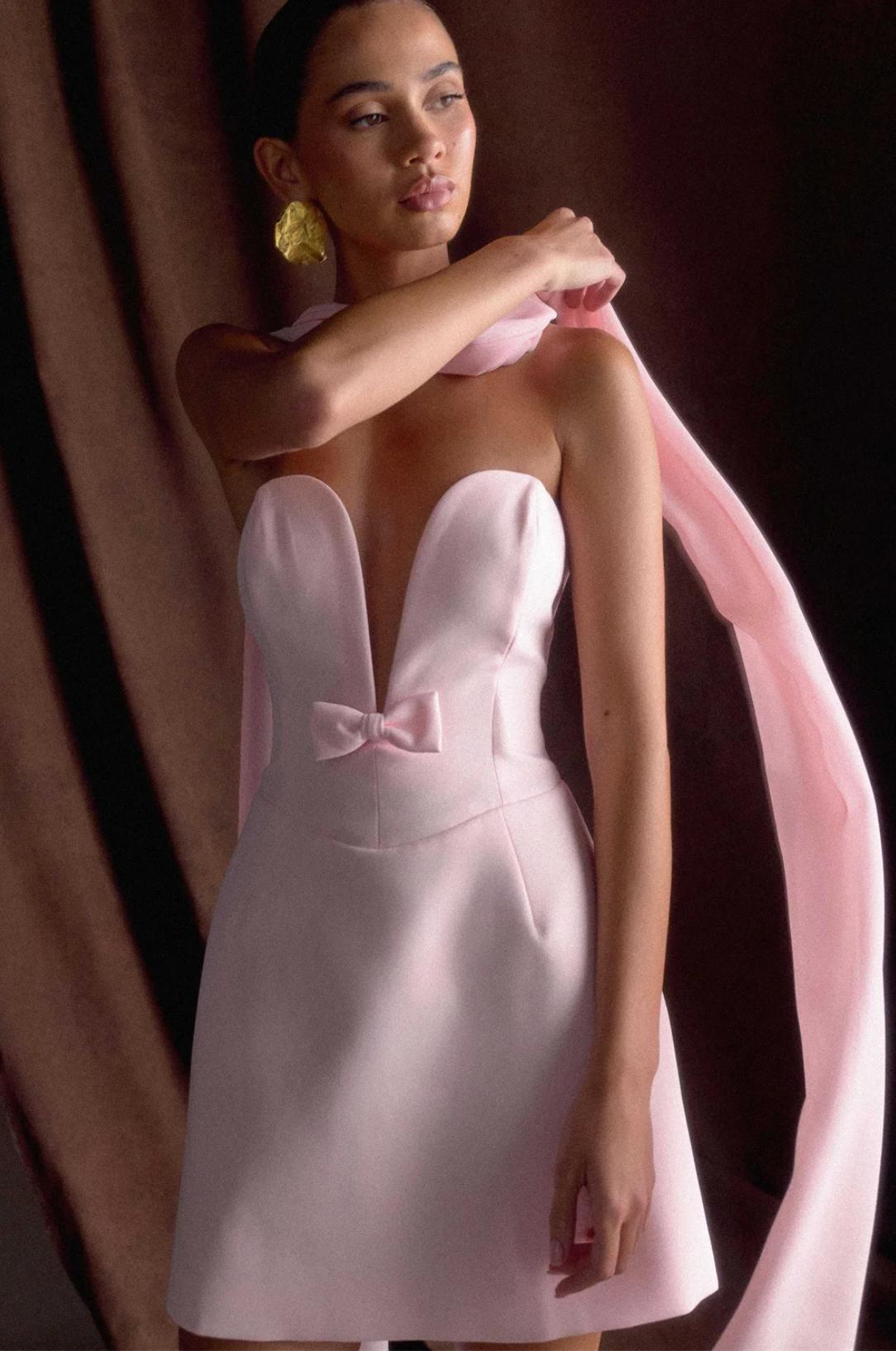 Model wearing a light pink A-line mini dress with a deep V-neckline, small front bow, and a matching removable shawl.