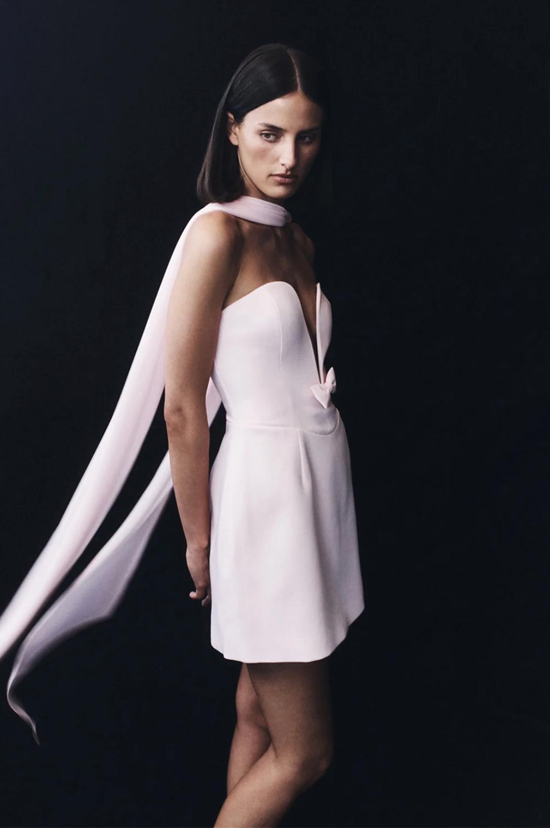 Model wearing a pale pink A-line mini dress with a deep V-neckline, small bow detail, and a flowing matching shawl.