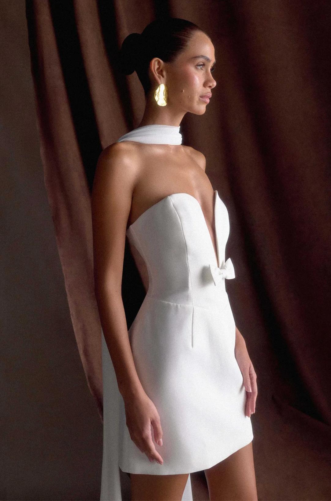 White mini dress with a deep V-neckline, small bow detail, and a removable shawl, crafted in a sleek, structured fabric.