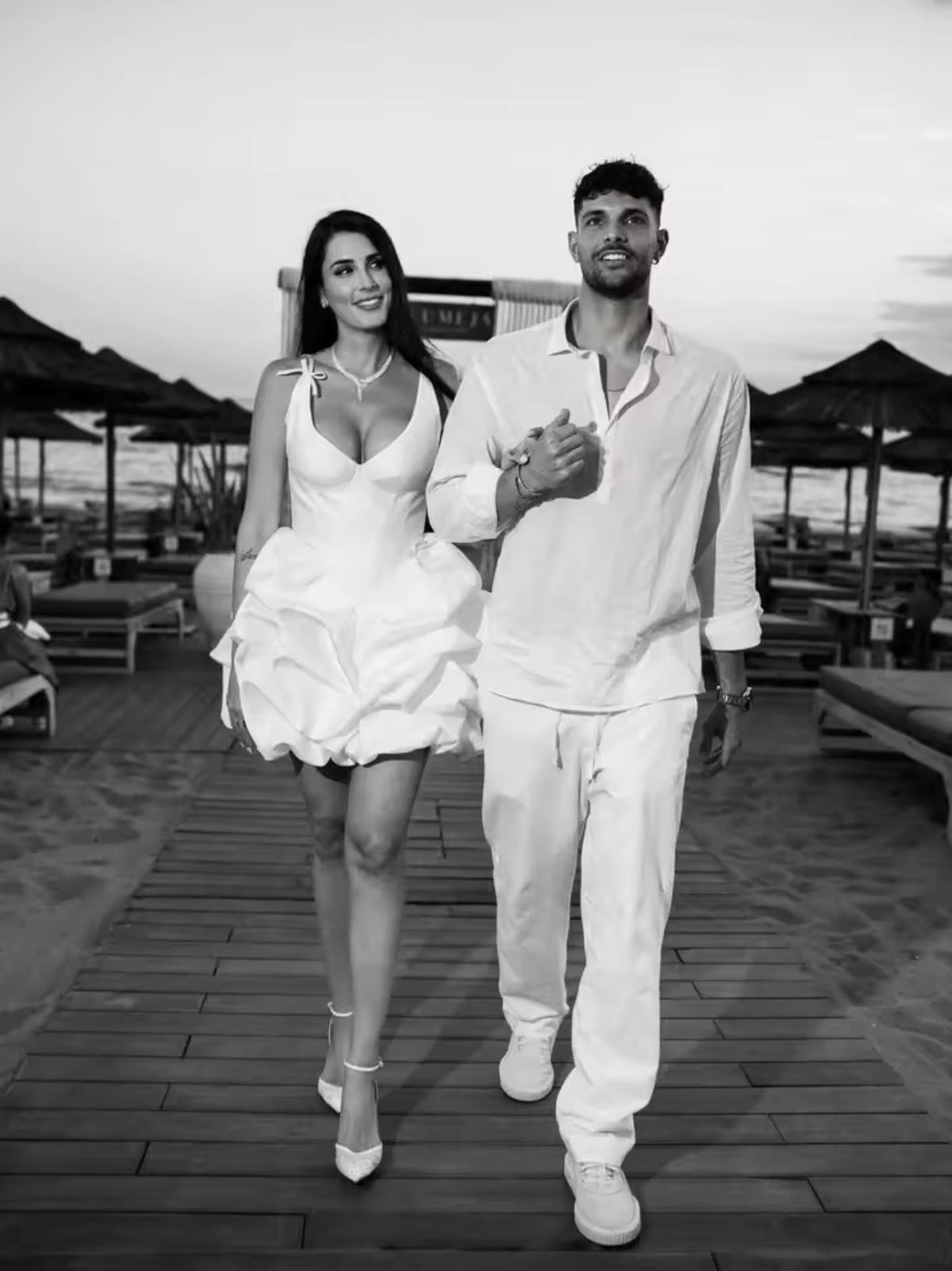 Woman in a white satin A-line mini dress with a plunging neckline and pleated skirt walking on a beach boardwalk.